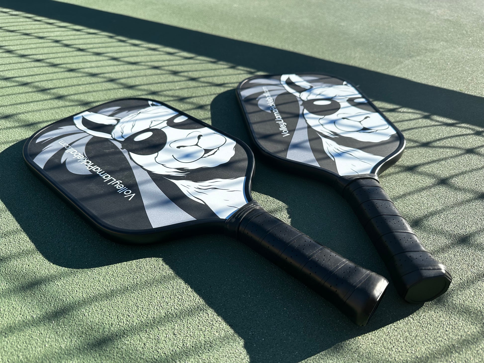 A pair of Holy Grail Pro Pickleball Paddles resting on the court in the shade of the net.